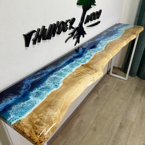 Puede incluir: Una mesa consola larga de madera con una tapa de resina blanca que presenta un diseño de olas de mar azules y blancas. La mesa tiene un marco de metal blanco y está sobre un suelo de madera. El texto "THUNDER WOOD" es visible en la pared detrás de la mesa.