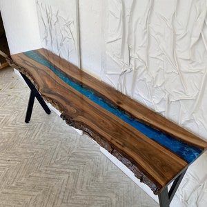 May include: A long console table featuring a natural live edge walnut wood slab with a central river inlay of vibrant blue epoxy resin. The table is supported by black metal legs and showcases the organic texture of the wood grain and bark.