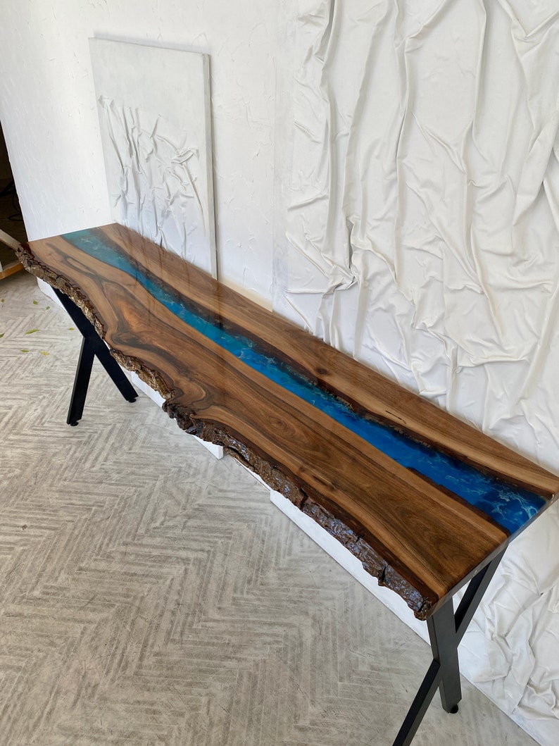 May include: A long console table featuring a natural live edge walnut wood slab with a central river inlay of vibrant blue epoxy resin. The table is supported by black metal legs and showcases the organic texture of the wood grain and bark.