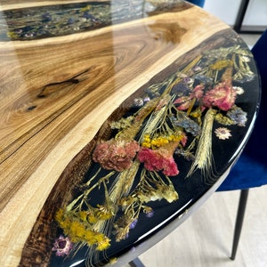 May include: Round wooden table with a live edge and a clear epoxy resin inlay. The resin section features dried flowers in shades of pink, yellow, and beige. The table has a dark metal base and is set against a blue velvet chair.