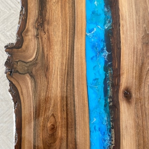 May include: A live edge wood slab table featuring a central river inlay made of vibrant blue resin. The natural wood grain displays rich brown tones with knots and organic curves, while the resin section shows swirling patterns of bright blue.