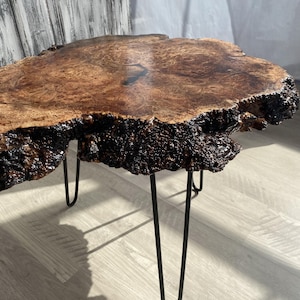 May include: A live edge coffee table with a rich, brown wood grain top. The table has a dark, textured edge and is supported by black hairpin legs. The wood grain has a glossy finish. The table is approximately 30 inches long.