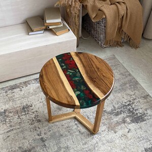 Puede incluir: Una mesa auxiliar redonda con un tablero de madera natural dividido por una incrustaci&oacute;n central de resina. La secci&oacute;n de resina contiene elementos florales rojos y follaje verde sobre un fondo oscuro. La mesa se apoya en tres patas de madera.