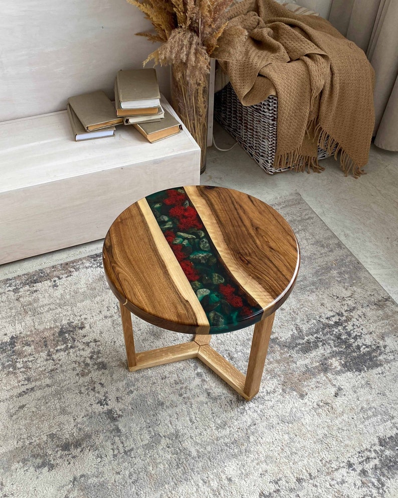 Puede incluir: Una mesa auxiliar redonda con un tablero de madera natural dividido por una incrustaci&oacute;n central de resina. La secci&oacute;n de resina contiene elementos florales rojos y follaje verde sobre un fondo oscuro. La mesa se apoya en tres patas de madera.