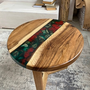 Puede incluir: Una mesa auxiliar redonda con dos tablones de madera natural unidos por una incrustaci&oacute;n central de resina verde transl&uacute;cida, incrustada con piedras grises lisas y texturas rojas vibrantes similares al musgo, sobre una base de madera.