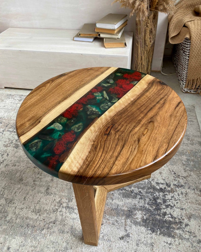 Puede incluir: Una mesa auxiliar redonda con dos tablones de madera natural unidos por una incrustaci&oacute;n central de resina verde transl&uacute;cida, incrustada con piedras grises lisas y texturas rojas vibrantes similares al musgo, sobre una base de madera.