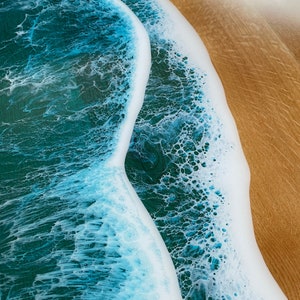 May include: Close-up of a resin art piece depicting a wave crashing on a sandy beach. The wave is a light blue color with white foam, and the sand is a light brown color.
