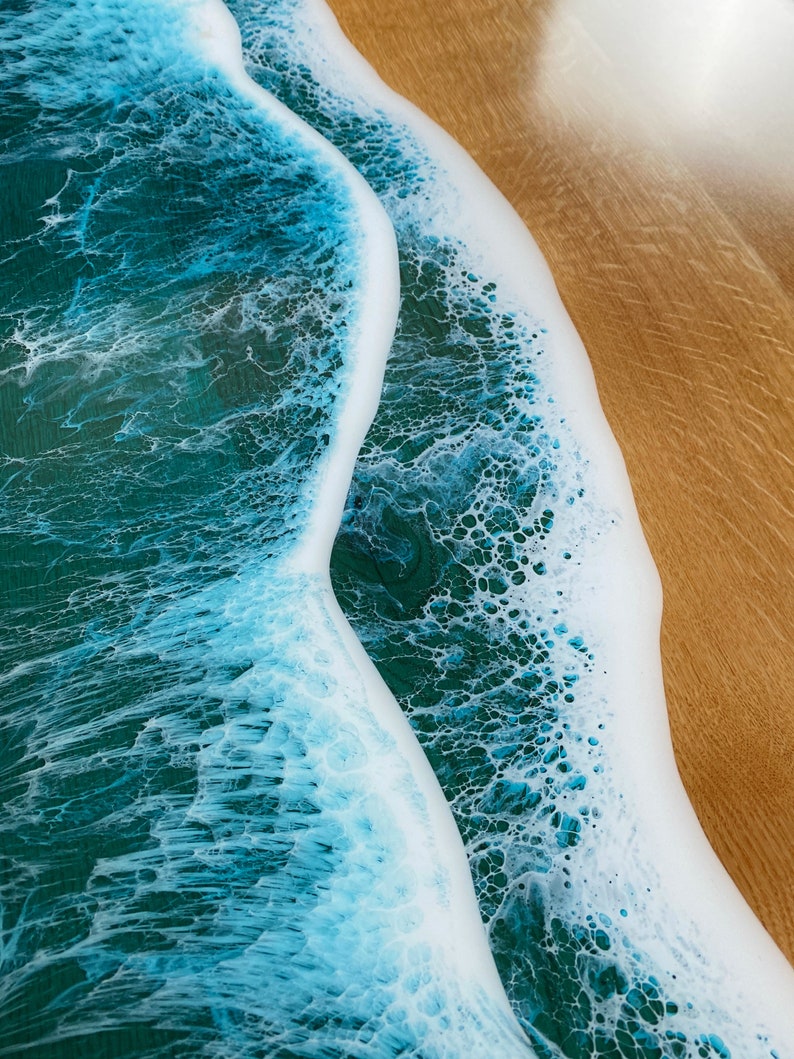 May include: Close-up of a resin art piece depicting a wave crashing on a sandy beach. The wave is a light blue color with white foam, and the sand is a light brown color.