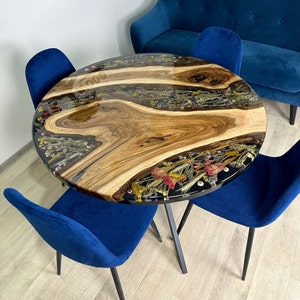 May include: Round wooden table with a clear resin top inlaid with dried flowers and wood grain. The table is supported by a black metal base and surrounded by four blue velvet chairs. The table's diameter is approximately 36 inches.