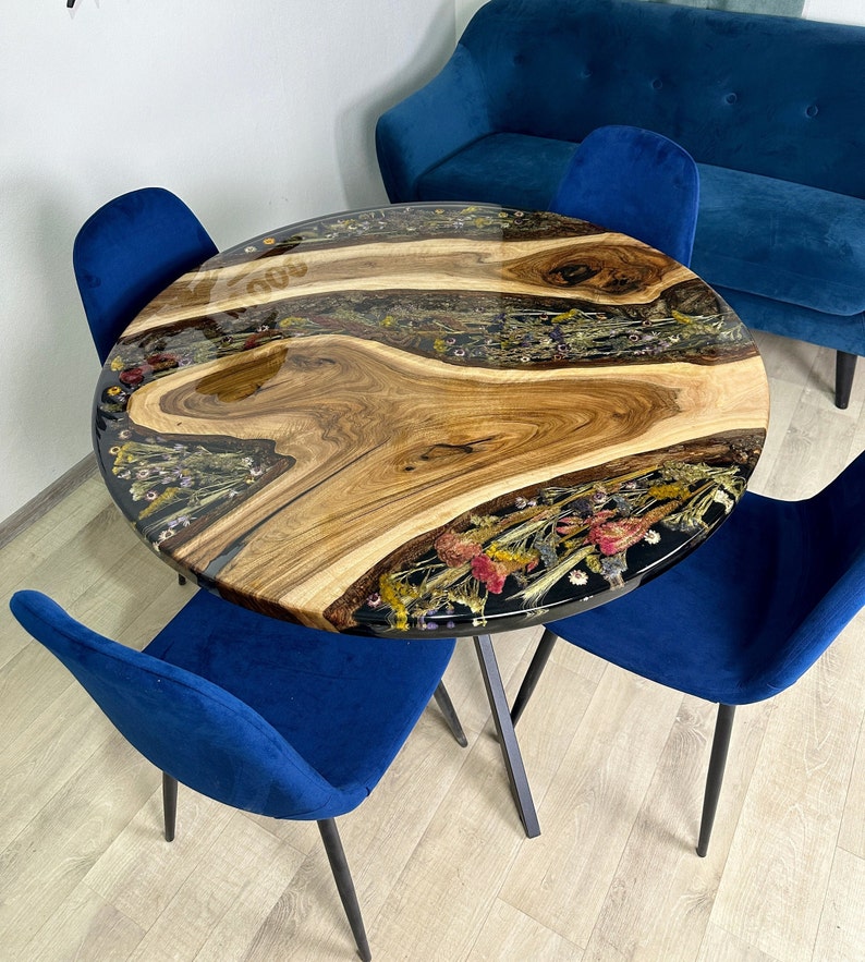 May include: Round wooden table with a clear resin top inlaid with dried flowers and wood grain. The table is supported by a black metal base and surrounded by four blue velvet chairs. The table's diameter is approximately 36 inches.