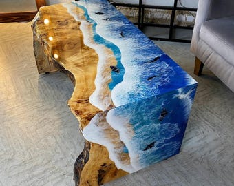 May include: A unique, handcrafted coffee table with a beach-themed design. The table features a live edge wood base with a resin top depicting ocean waves and sand. The colors include shades of blue, white, and brown. The table is approximately 48 inches long.
