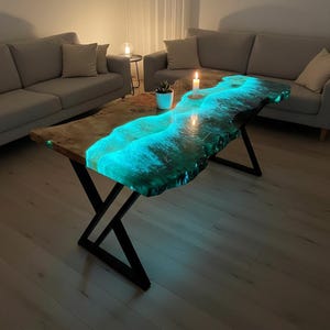 May include: A unique coffee table with a live edge design. The table features a glowing blue resin river that runs through the center, creating a striking visual effect. The table is supported by black metal legs.