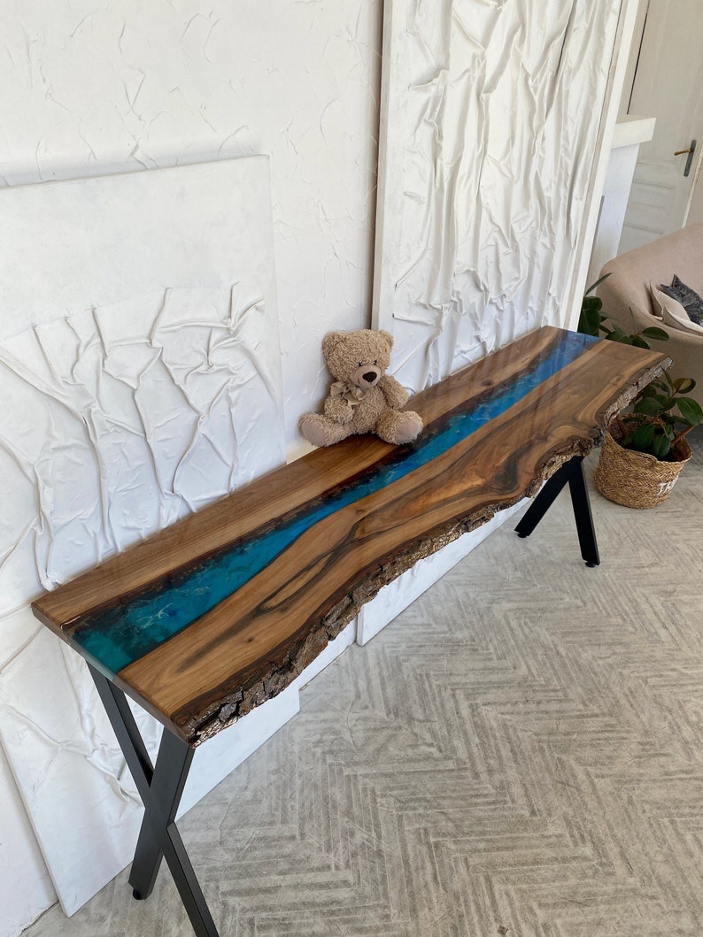 May include: A long console table featuring a natural live edge wood slab with a central river of translucent blue epoxy resin. The table is supported by black metal X-shaped legs and displays a prominent wood grain with dark brown tones.