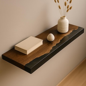 May include: A floating wooden shelf with a dark wood grain finish and a black border. A small white vase with dried flowers, a beige book, and a white egg-shaped object are on the shelf. The shelf is mounted on a neutral-colored wall.