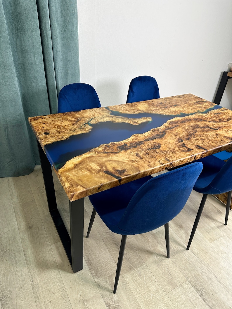 May include: A rectangular wooden dining table with a blue epoxy river design, supported by black metal legs. Four blue velvet chairs surround the table. The table's wood grain is visible, and the epoxy creates a contrasting blue river effect.