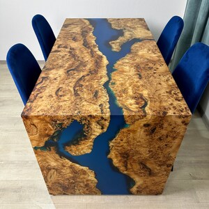 May include: A rectangular wooden table with a blue river-like epoxy resin inlay. The table has a natural wood grain pattern and is surrounded by four blue velvet chairs. The table's dimensions are approximately 72 inches long, 36 inches wide, and 30 inches high.