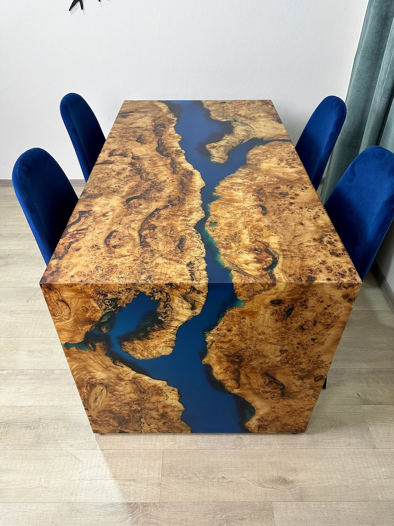 May include: A rectangular wooden table with a blue river-like epoxy resin inlay. The table has a natural wood grain pattern and is surrounded by four blue velvet chairs. The table's dimensions are approximately 72 inches long, 36 inches wide, and 30 inches high.