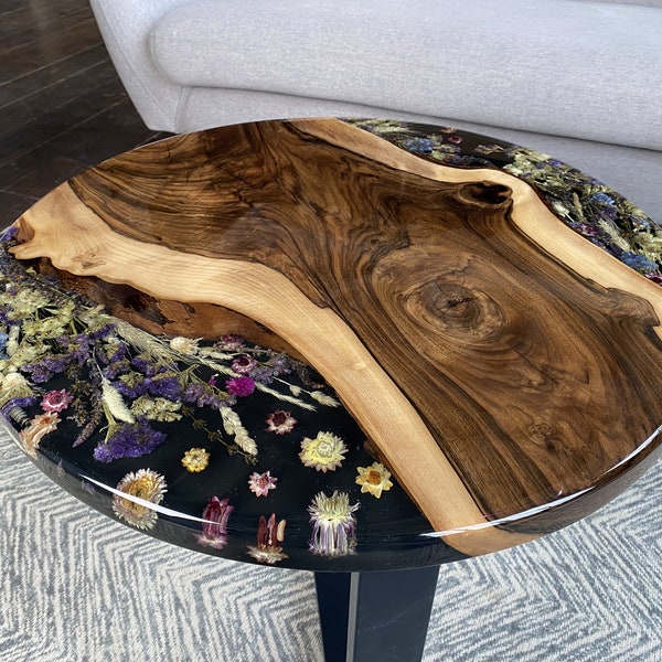 Può includere: Un tavolino rotondo caratterizzato da una lastra di legno naturale lucido con venature e nodi prominenti, combinato con una sezione in resina nera con fiori secchi incorporati nei toni del viola, rosa, giallo e verde, su una base scura.