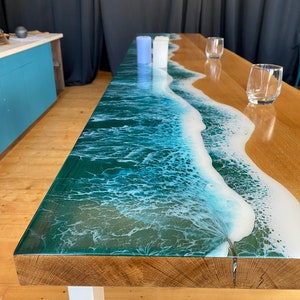 May include: A wooden table with a clear epoxy resin top featuring a blue and white ocean wave design. The table has a white metal base and is set with two clear glasses and three candles.