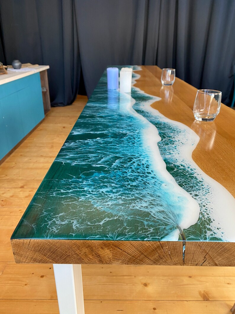 May include: A wooden table with a clear epoxy resin top featuring a blue and white ocean wave design. The table has a white metal base and is set with two clear glasses and three candles.