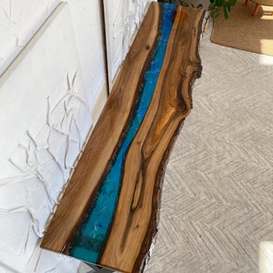 May include: A long, narrow console table featuring a natural live-edge walnut wood slab with a prominent, vibrant blue resin river inlay running through the center. The table is supported by a dark metal base and has a smooth, polished finish.