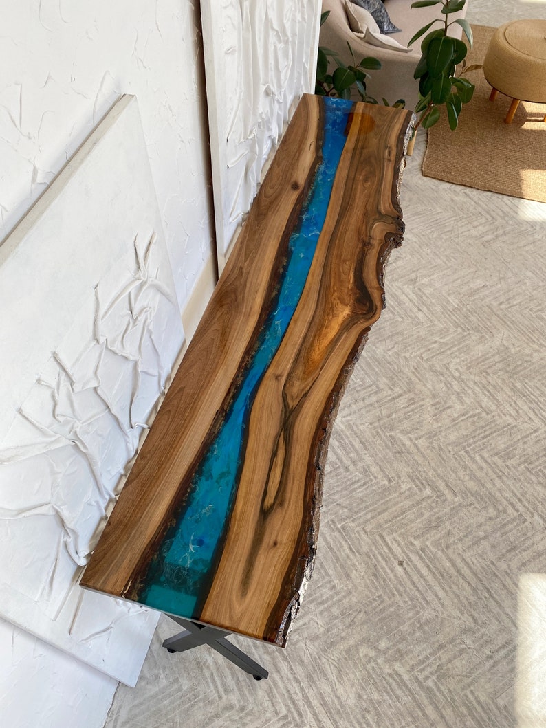 May include: A long, narrow console table featuring a natural live-edge walnut wood slab with a prominent, vibrant blue resin river inlay running through the center. The table is supported by a dark metal base and has a smooth, polished finish.
