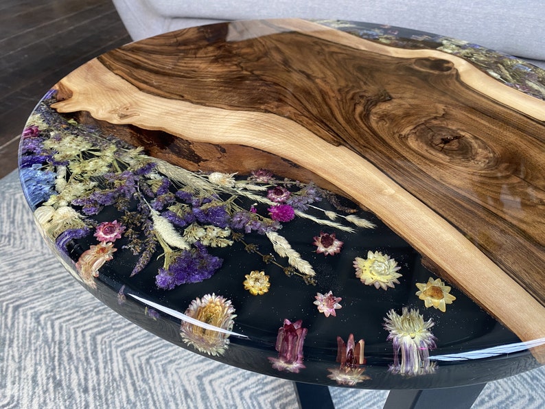 K&ouml;nnte beinhalten: Ovaler Couchtisch mit einem Design mit lebendiger Kante. Der Tisch hat eine dunkle Holzmaserung und einen Abschnitt aus schwarzem Harz mit getrockneten Blumen in Lila, Rosa und Gelb. Der Tisch hat eine gl&auml;nzende Oberfl&auml;che.