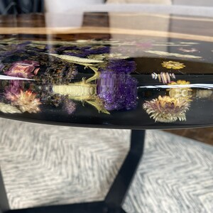 K&ouml;nnte beinhalten: Runder Couchtisch mit einer schwarzen Harzplatte, in die getrocknete Blumen in Lila, Gelb und Wei&szlig; eingelegt sind. Der Tisch hat einen dunklen Holzrand und schwarze Metallbeine, was eine moderne &Auml;sthetik schafft.
