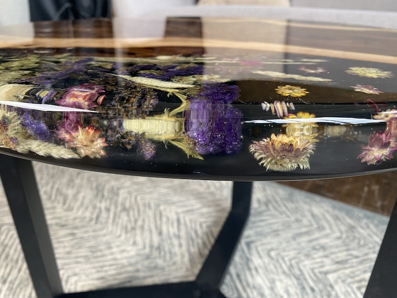 K&ouml;nnte beinhalten: Runder Couchtisch mit einer schwarzen Harzplatte, in die getrocknete Blumen in Lila, Gelb und Wei&szlig; eingelegt sind. Der Tisch hat einen dunklen Holzrand und schwarze Metallbeine, was eine moderne &Auml;sthetik schafft.