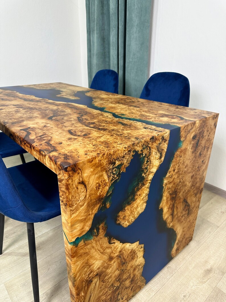 May include: A handcrafted wooden dining table with a unique design. The table features a natural wood grain surface with a river of deep blue resin flowing through the center. Two blue chairs are visible in the background.