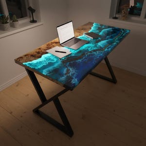 May include: A rectangular desk featuring a natural wood slab with a live edge and a glowing blue resin inlay that mimics ocean waves. The desk is supported by a black metal frame with a Z-shaped base. A laptop, a notebook, and a pen sit on the surface.