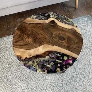 K&ouml;nnte beinhalten: Runder Couchtisch aus Holz mit einer Baumkante und einem schwarzen Harzeinlegearbeiten, gef&uuml;llt mit getrockneten Blumen. Der Tisch hat eine nat&uuml;rliche Holzmaserung und schwarze Metallbeine. Der Tisch hat einen Durchmesser von ca. 91 cm.