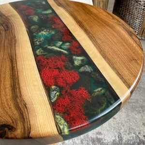 Puede incluir: Una mesa redonda de madera con una incrustaci&oacute;n central de resina transparente. La incrustaci&oacute;n contiene una colecci&oacute;n de piedras grises lisas y musgo preservado de color rojo vibrante, creando un aspecto natural en la superficie.