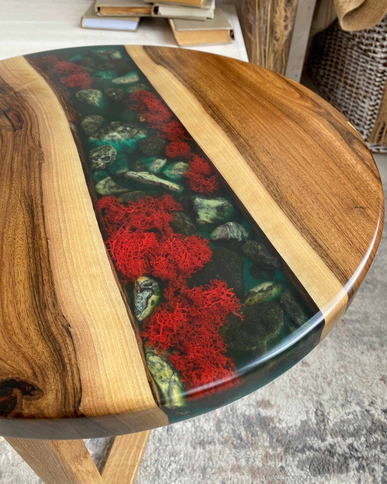 Puede incluir: Una mesa redonda de madera con una incrustaci&oacute;n central de resina transparente. La incrustaci&oacute;n contiene una colecci&oacute;n de piedras grises lisas y musgo preservado de color rojo vibrante, creando un aspecto natural en la superficie.