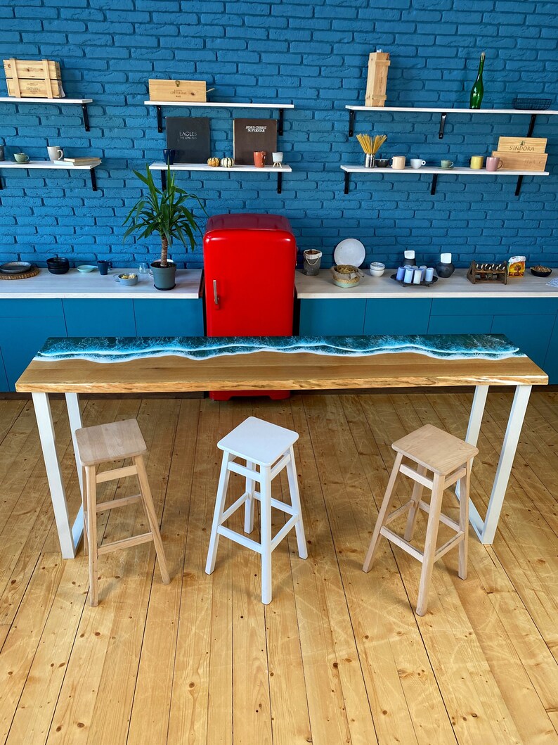 May include: A long wooden table with a blue epoxy resin wave design. The table has white metal legs and is set with three wooden stools. Two of the stools are natural wood and one is white.