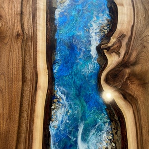 May include: A close-up of a wooden table with a blue and white epoxy river running through the center. The epoxy has a swirling pattern and white accents that resemble waves.