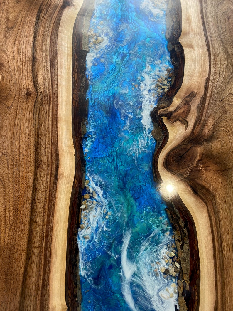 May include: A close-up of a wooden table with a blue and white epoxy river running through the center. The epoxy has a swirling pattern and white accents that resemble waves.