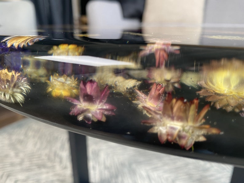 K&ouml;nnte beinhalten: Nahaufnahme eines schwarzen Harztisches mit eingebetteten getrockneten Blumen. Die Blumen sind in Gelb-, Rosa- und Wei&szlig;t&ouml;nen gehalten. Der Rand des Tisches ist sichtbar und zeigt eine glatte, gl&auml;nzende Oberfl&auml;che.