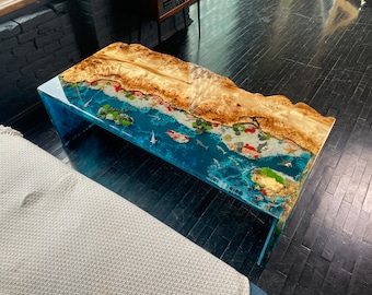 May include: A rectangular coffee table with a unique design. The table features a wooden top with a natural edge, combined with a blue resin base that incorporates a miniature ocean scene with fish and coral. The table's dimensions are approximately 48 inches long.