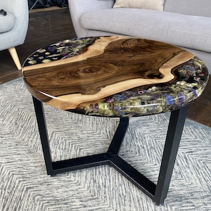K&ouml;nnte beinhalten: Runder Couchtisch mit einer Tischplatte aus Holz mit nat&uuml;rlicher Kante. Der Tisch hat eine dunkle Holzmaserung in der Mitte, umgeben von klarem Harz mit getrockneten Blumen. Er hat ein schwarzes Metallgestell. Durchmesser ca. 76 cm.