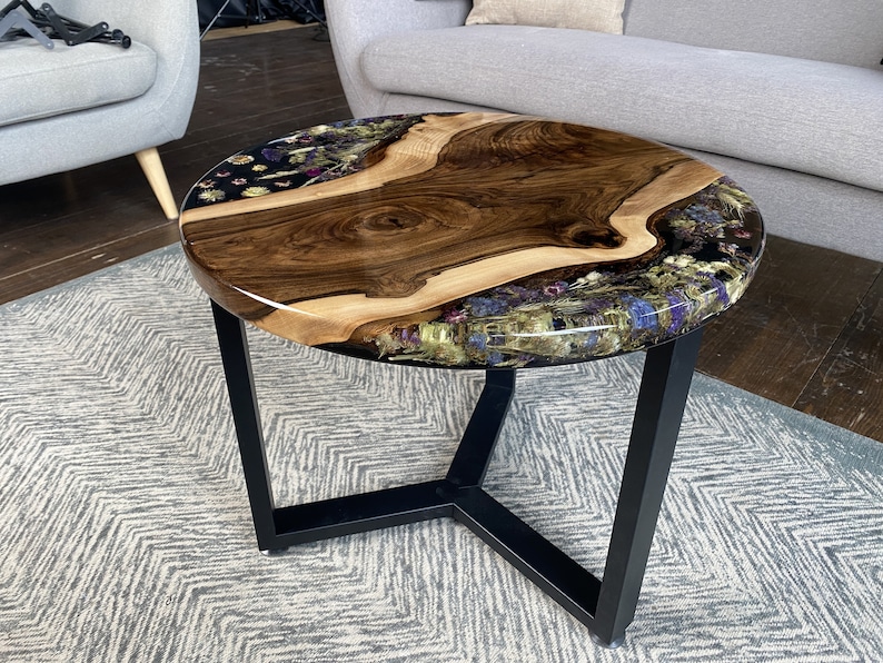 K&ouml;nnte beinhalten: Runder Couchtisch mit einer Tischplatte aus Holz mit nat&uuml;rlicher Kante. Der Tisch hat eine dunkle Holzmaserung in der Mitte, umgeben von klarem Harz mit getrockneten Blumen. Er hat ein schwarzes Metallgestell. Durchmesser ca. 76 cm.