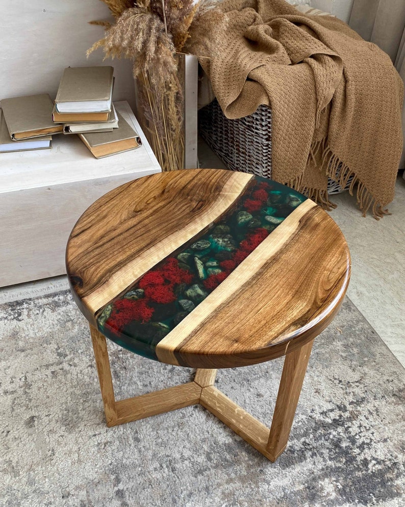 Puede incluir: Una mesa auxiliar redonda con un tablero de madera de veta natural que presenta una incrustaci&oacute;n central de resina verde oscuro, con piedras grises lisas y texturas rojas similares al musgo, sostenida por tres patas de madera clara.