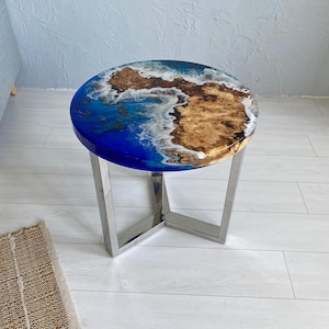 May include: A round side table featuring a live edge wood burl top embedded in blue resin that mimics ocean waves with white foam detailing. The table is supported by a three-legged base made of polished silver-colored metal with a triangular frame.