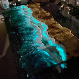 Puede incluir: Primer plano de una mesa de madera con un río de resina epoxi azul y verde. La resina es translúcida y brilla en la oscuridad, creando un efecto de onda.