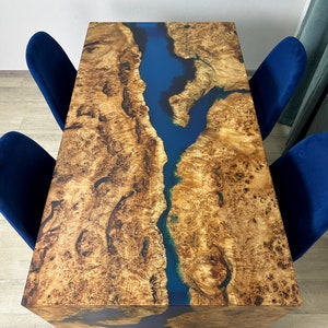 May include: A rectangular wooden table with a blue river-like epoxy resin inlay. The table's surface has a natural wood grain pattern in shades of brown and tan. Four blue velvet chairs surround the table, creating a modern dining set.