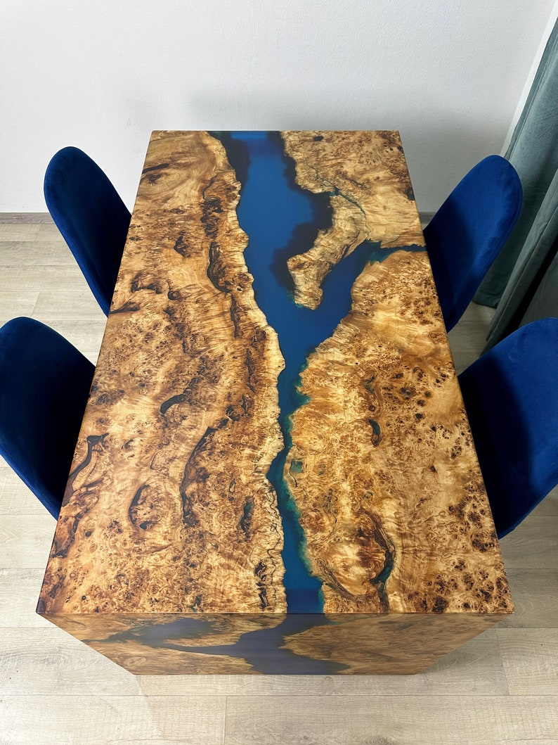 May include: A rectangular wooden table with a blue river-like epoxy resin inlay. The table's surface has a natural wood grain pattern in shades of brown and tan. Four blue velvet chairs surround the table, creating a modern dining set.