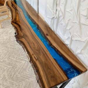 May include: A long rectangular console table featuring a live edge walnut wood slab with a central river inlay made of vibrant blue epoxy resin. The table has a polished, glossy finish and is supported by a black metal X-shaped base frame.