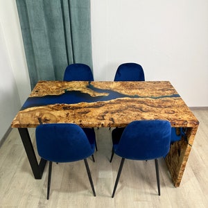 May include: A wooden dining table with a blue epoxy river design and four blue velvet chairs. The table has a natural wood grain finish and black metal legs. The chairs have black metal legs.