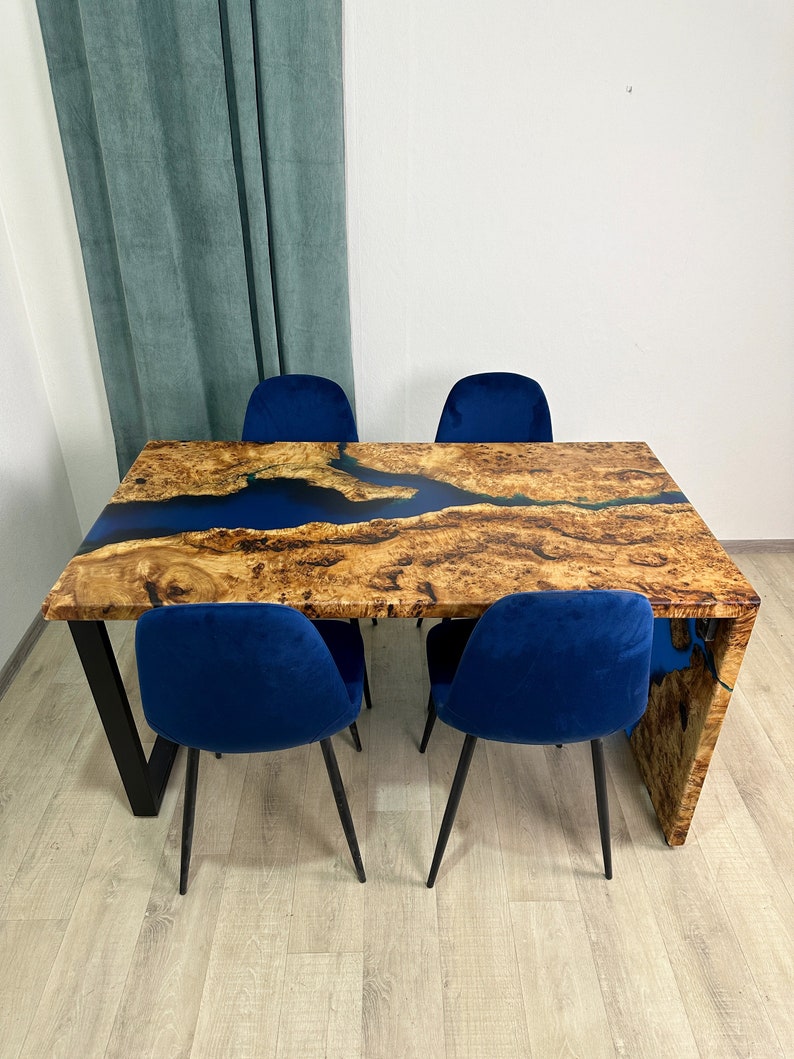 May include: A wooden dining table with a blue epoxy river design and four blue velvet chairs. The table has a natural wood grain finish and black metal legs. The chairs have black metal legs.