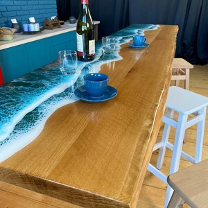 May include: A wooden table with a white epoxy resin top featuring a blue ocean wave design. The table has a white base and is surrounded by wooden and white stools.